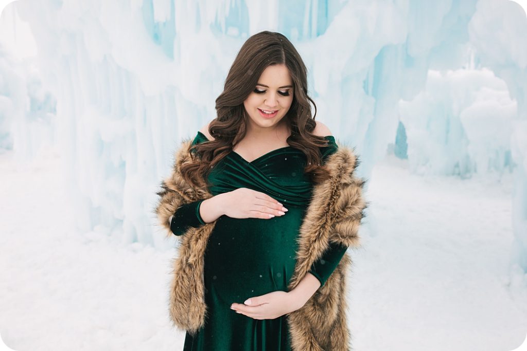 mom in velvet maternity gown with fur poses in Ice Castles display for maternity session