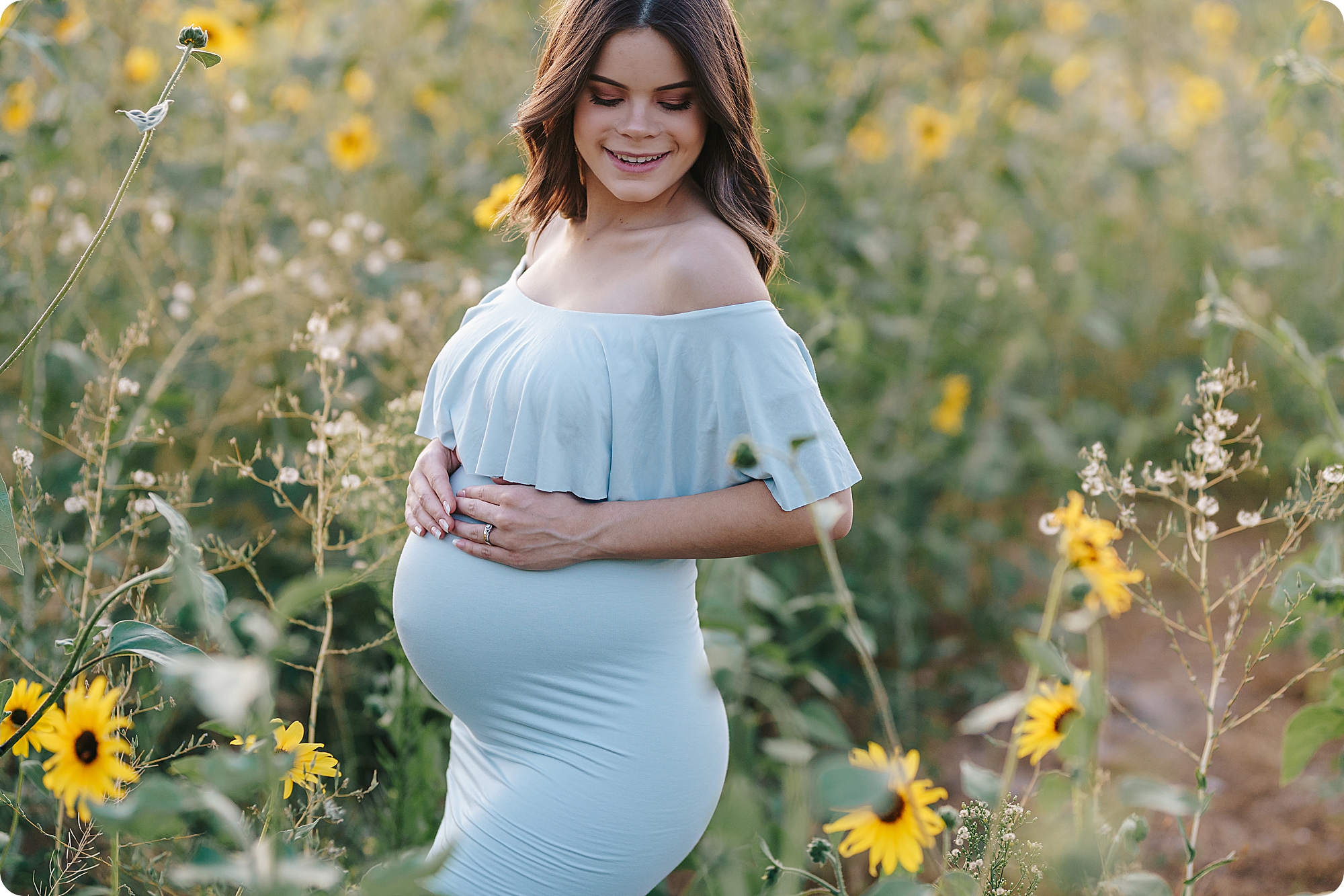 mom holds belly wearing light blue gown in sunflower field