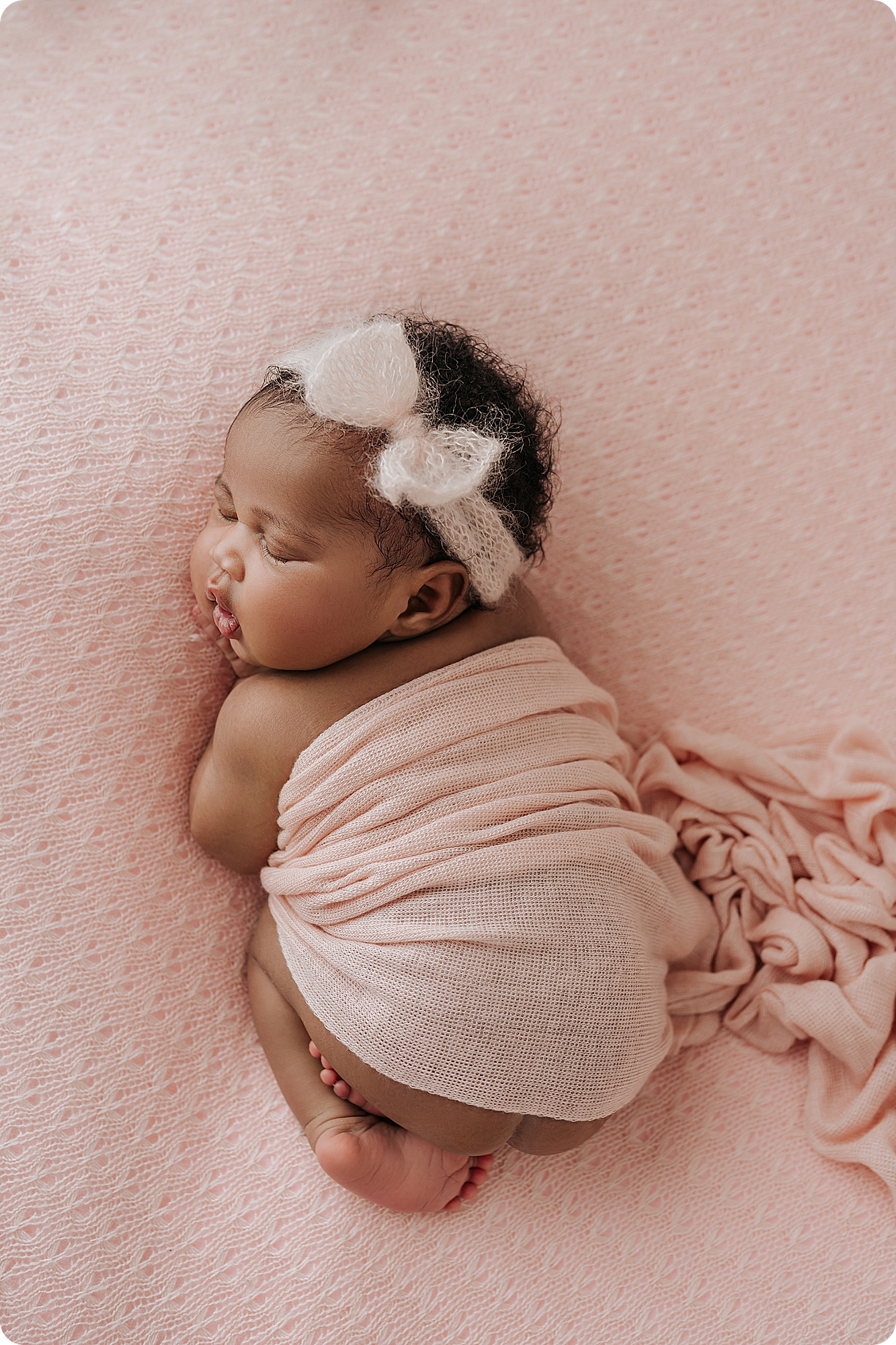 newborn baby girl with bow in hair sleeps on pastel pink blanket
