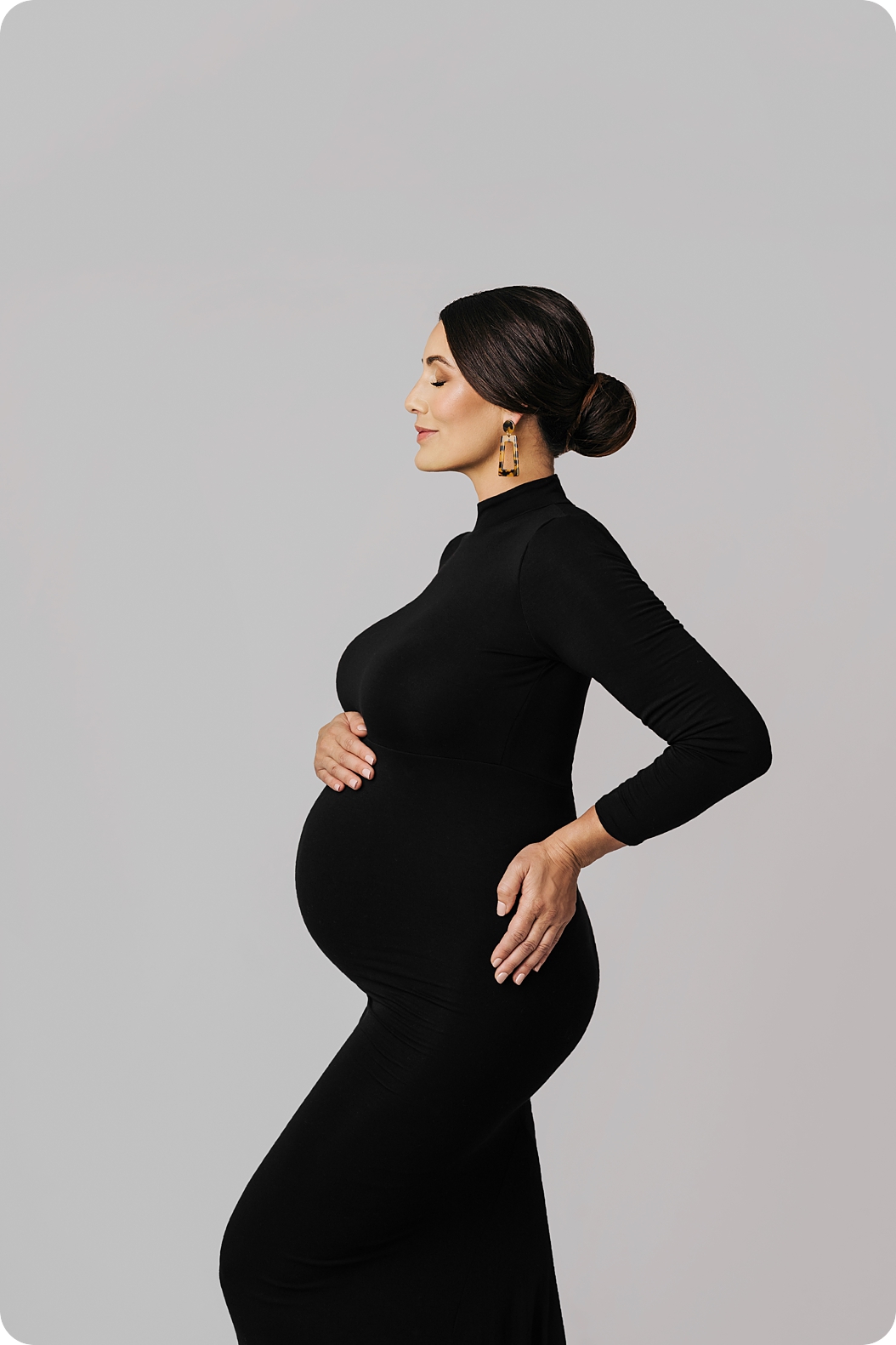 mom stands with eyes closed and hair in bun with leopard inspired earrings holding baby belly during Classic Studio Maternity Portraits