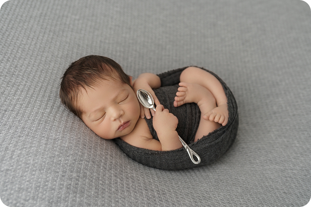 baby boy holds spoon while curled up during simple newborn portraits with Beka Price Photography