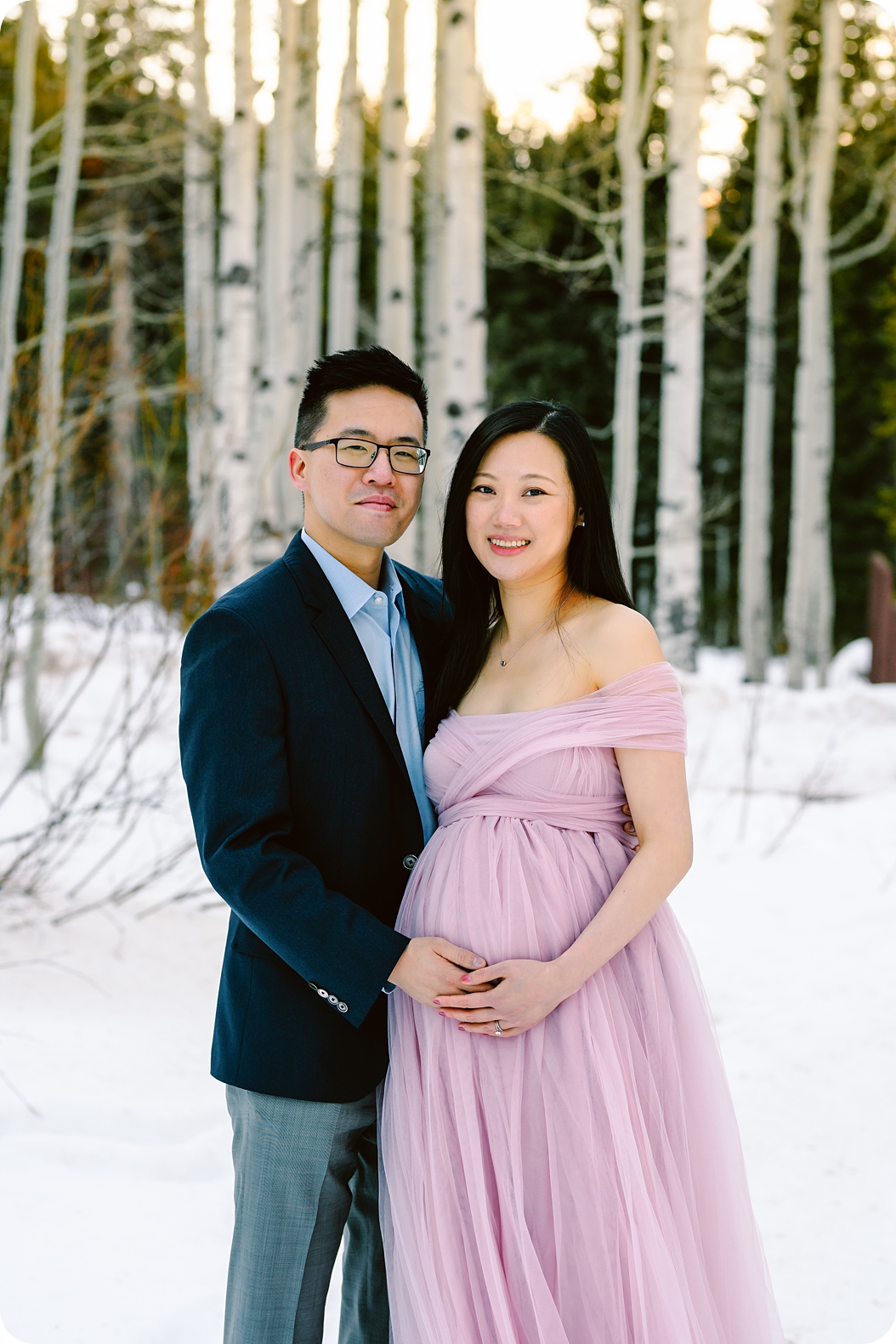 mom in pink maternity gown poses next to dad during photos to celebrate pregnancy in the snow