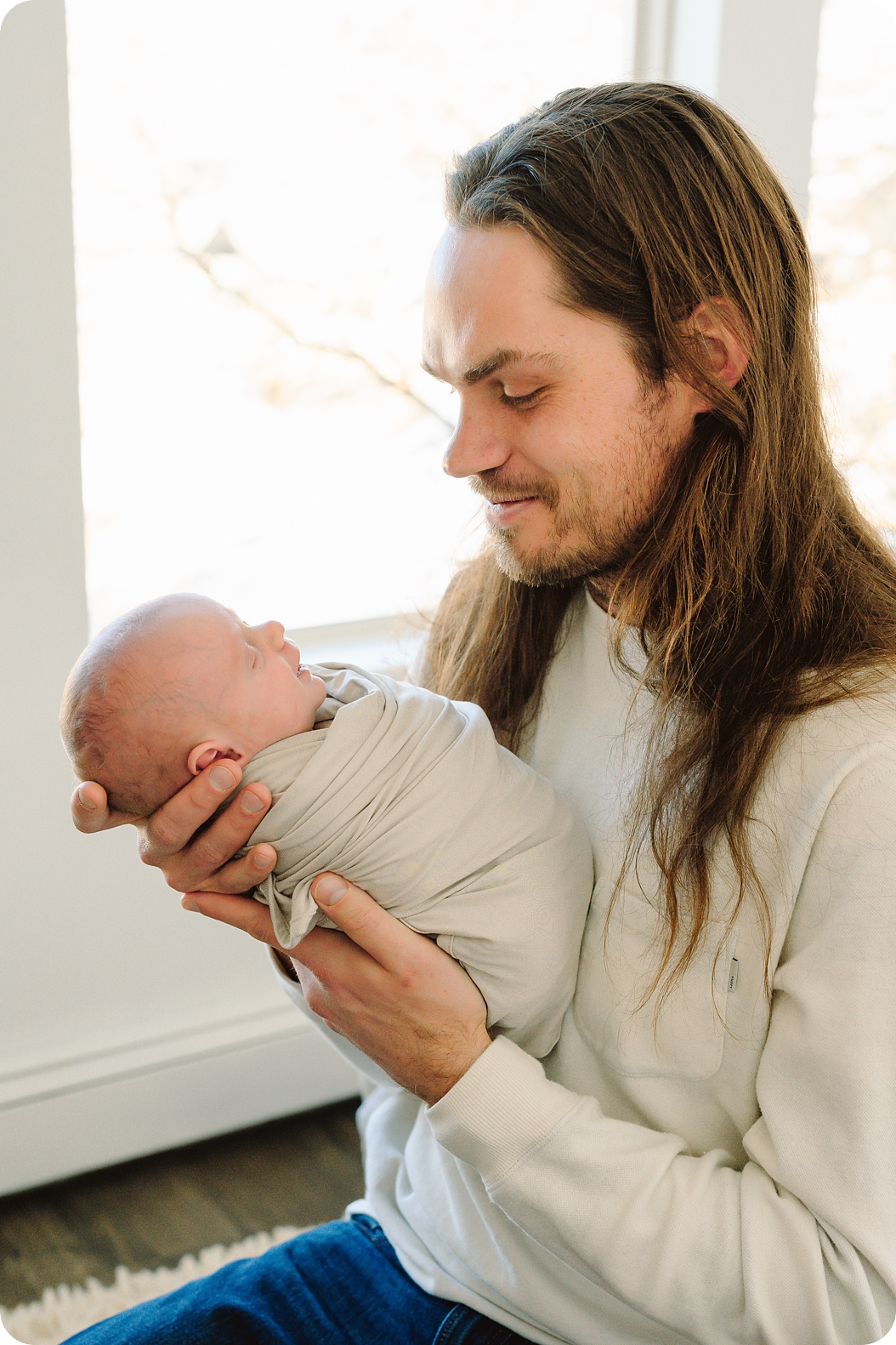 dad with long hair looks down at newborn baby girl during lifestyle photos at home 
