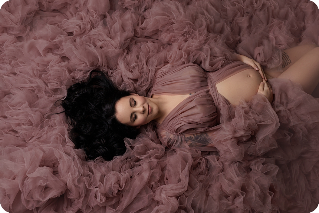 mother in mauve dress lays back on fluff holding belly in Utah studio 