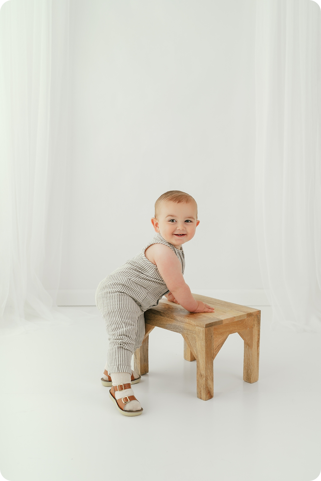 baby in striped romper leans against wooden stool during birthday photos