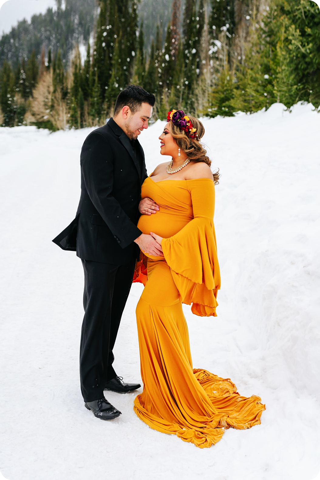 parents pose in the snow holding mom's belly in mustard gown during Utah maternity photos 