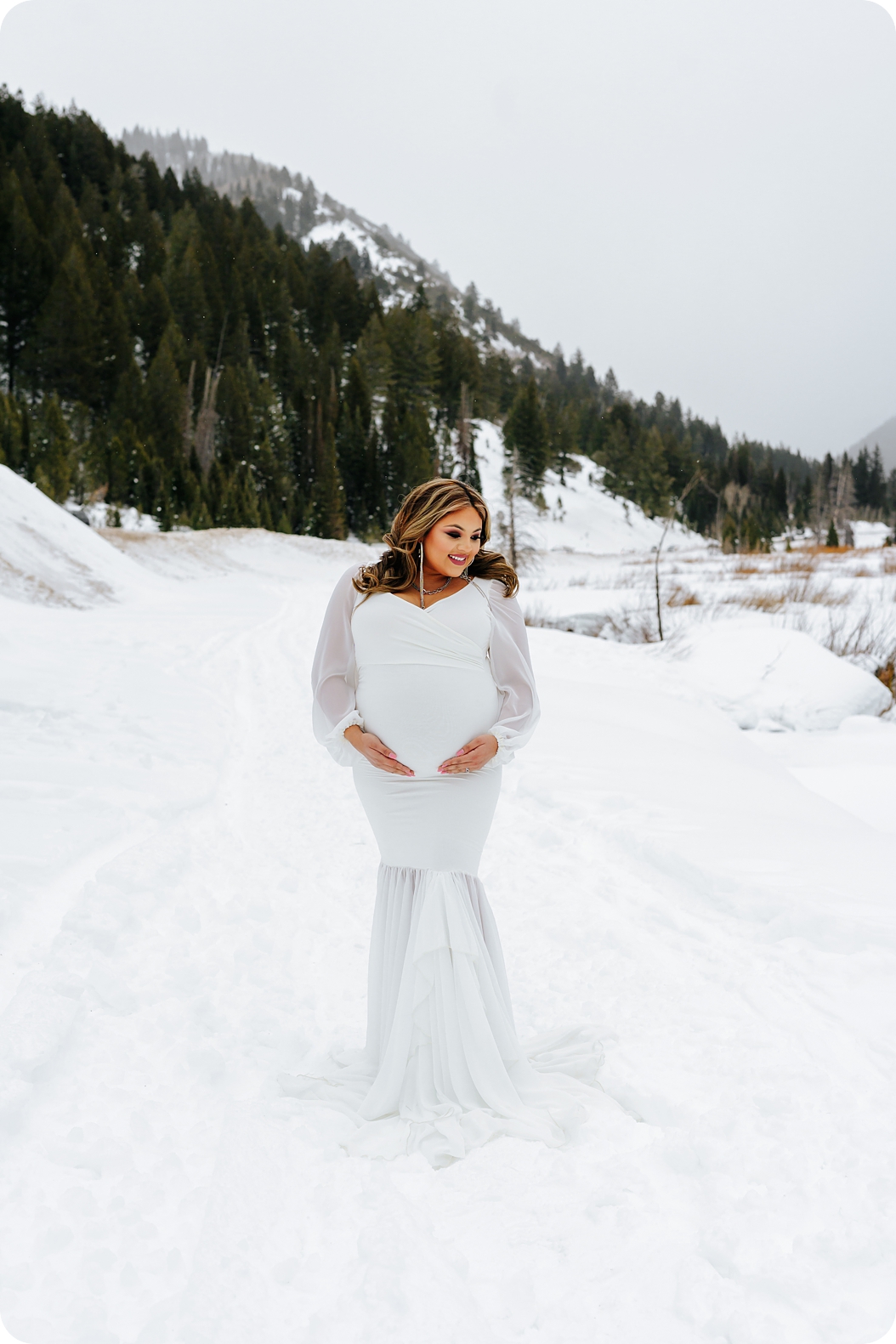 mom in white maternity gown poses in the snow during Dramatic Winter Maternity Portraits