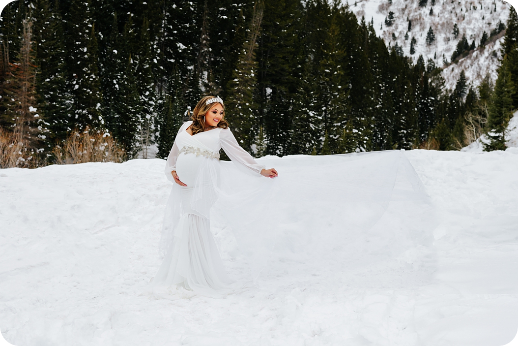 mom in white gown looks over shoulder in the snow during maternity photos 