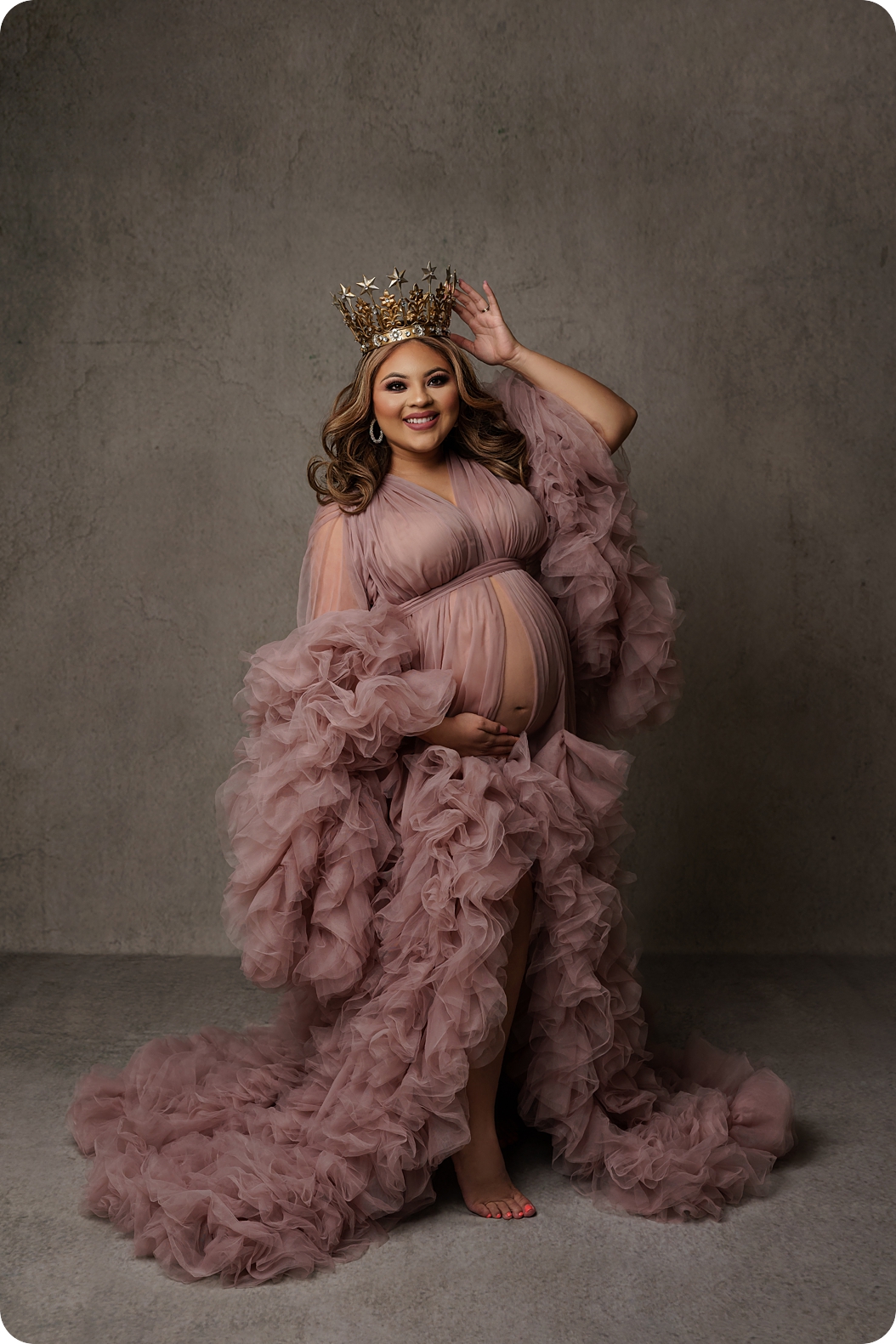 expecting mother holds belly during maternity portraits in blush pink maternity gown with crown in studio 