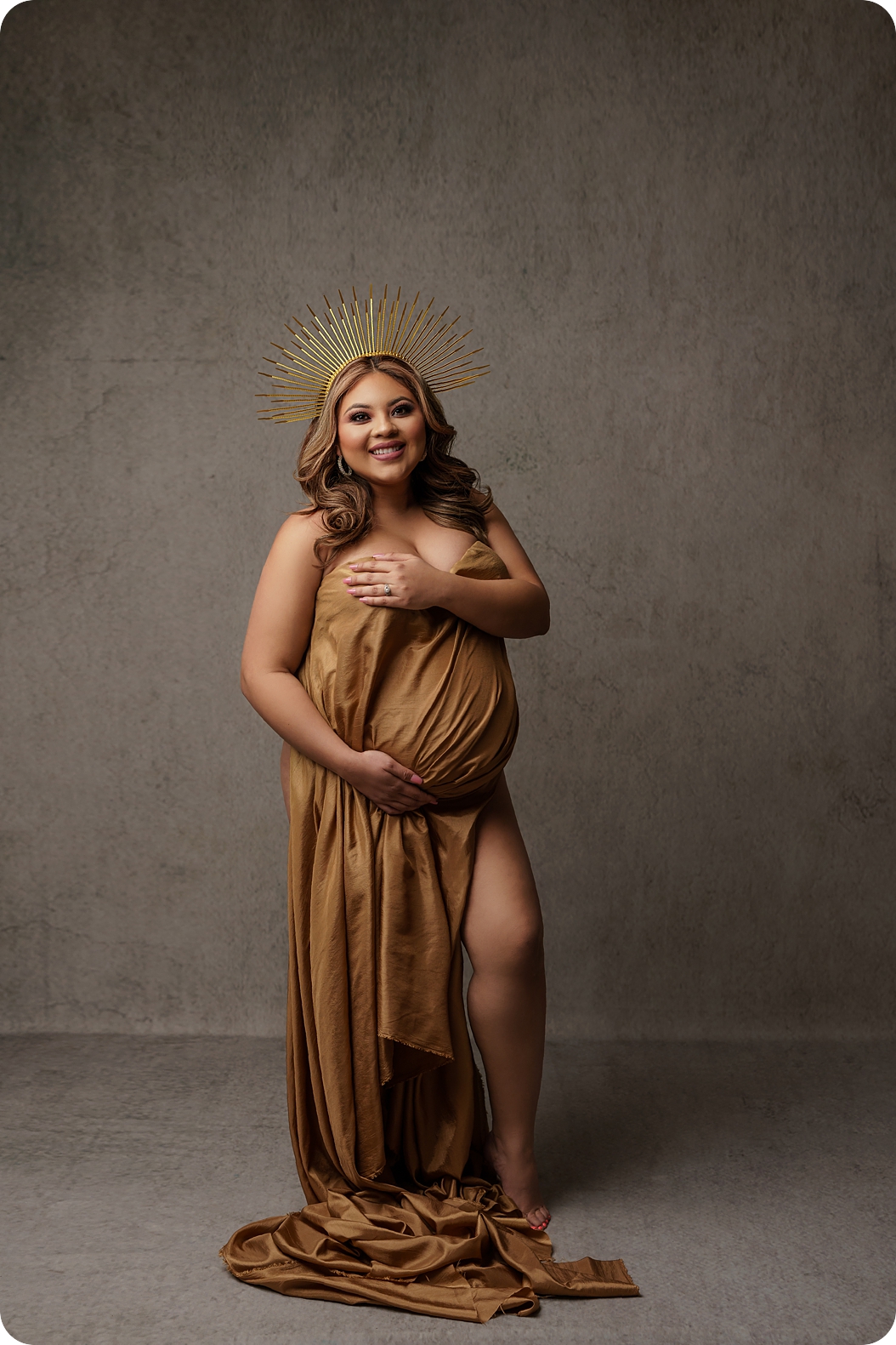 mother holds gold wrap over stomach wearing gold crown during maternity photos 