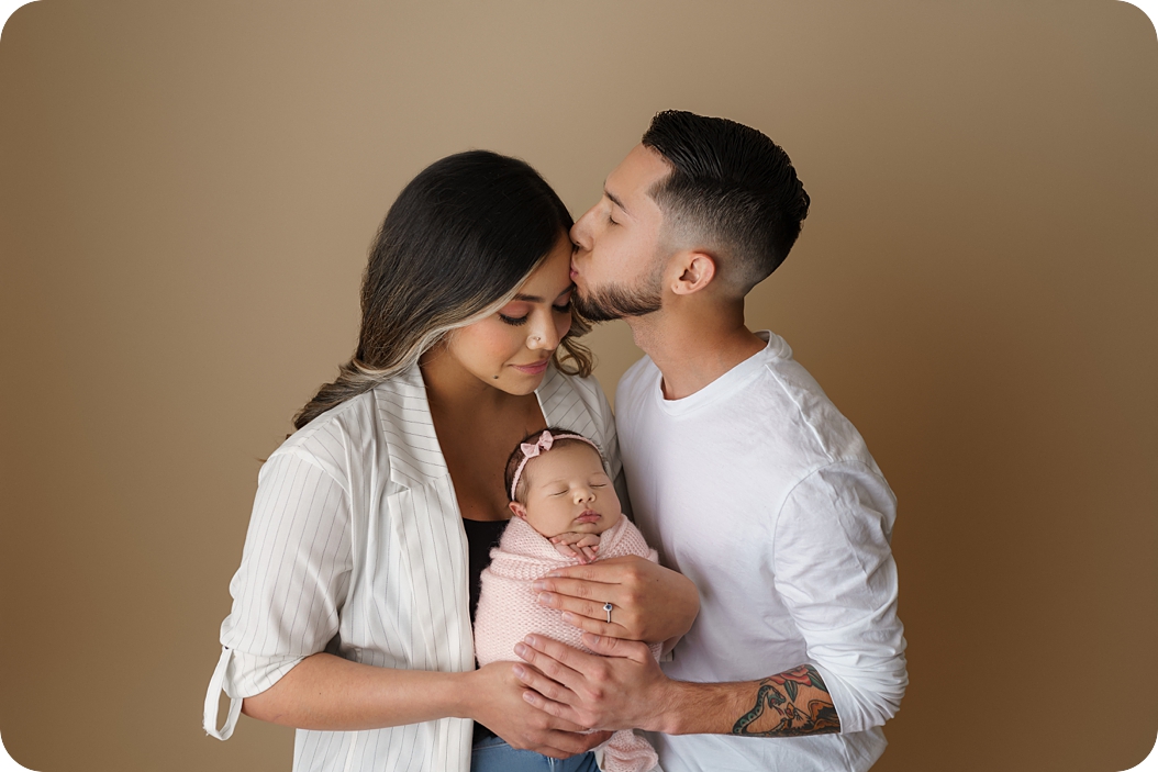 dad kisses mom's forehead while she holds baby girl during pastel newborn portraits 
