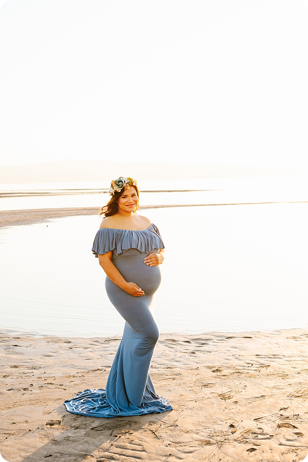 woman in light blue gown poses in sand during waterfront maternity portraits 