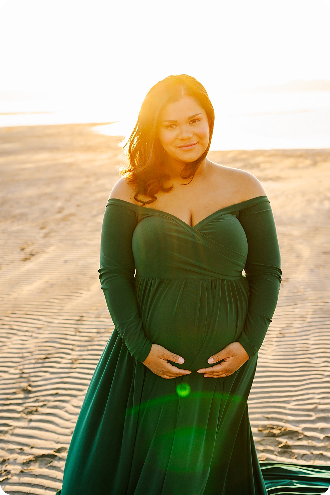 woman holds belly in green gown at sunset during waterfront maternity portraits 