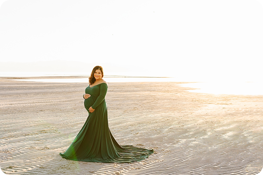 mother in green gown holds belly standing on sand during waterfront maternity portraits 