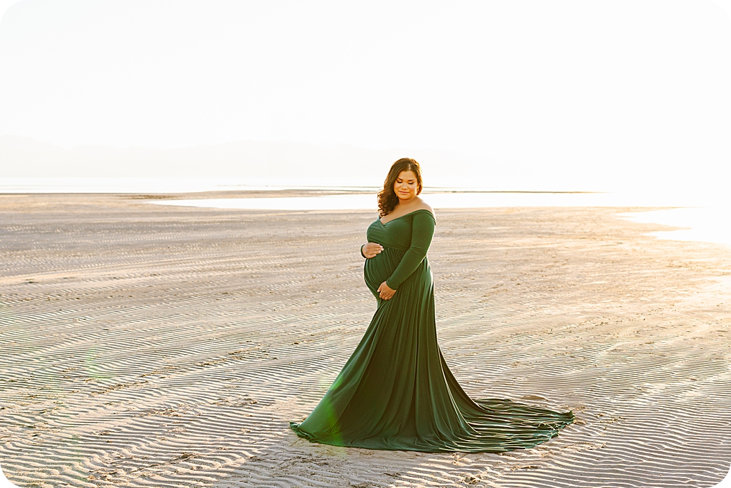 woman in green gown stands on sand holding belly