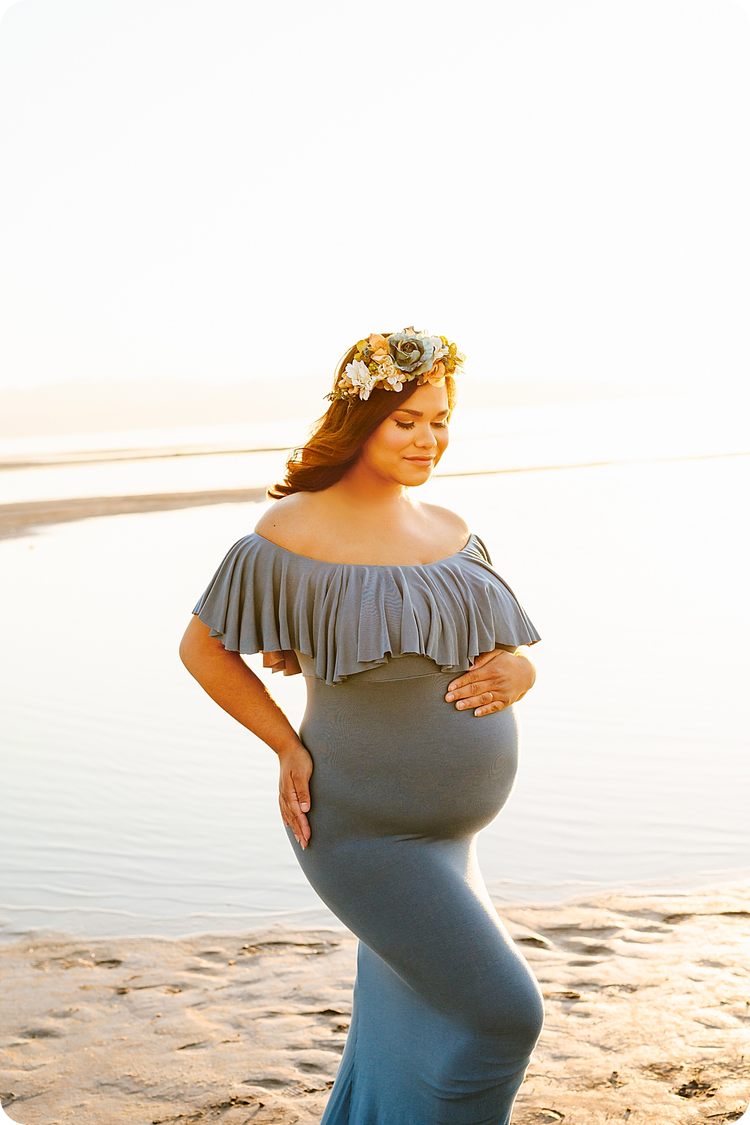 expecting mother in blue gown with floral crown holds baby belly during waterfront maternity portraits 