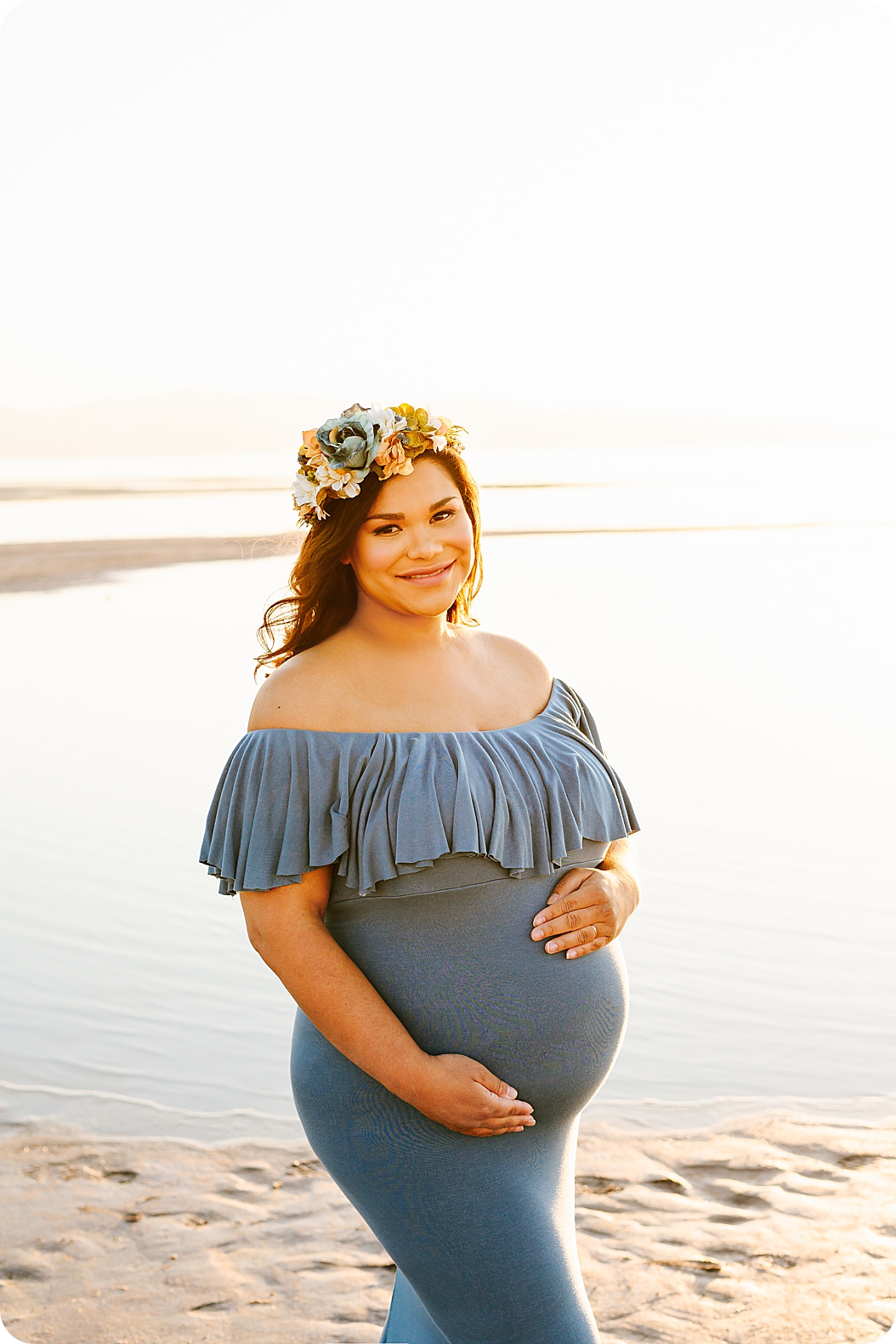 mom holds belly during waterfront maternity portraits in blue gown with floral crown