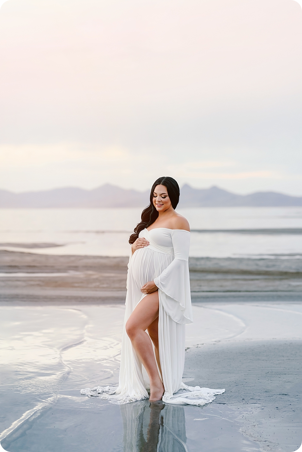 woman holds belly in white maternity gown along lake