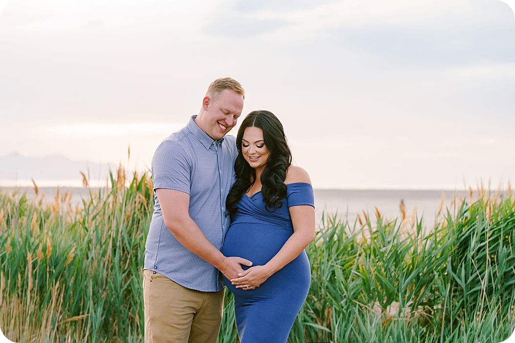 parents-to-be look at belly during maternity photos by Utah lake 