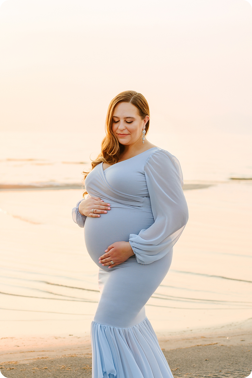 mom in long sleeve blue gown holds belly during sunset maternity portraits on the lake