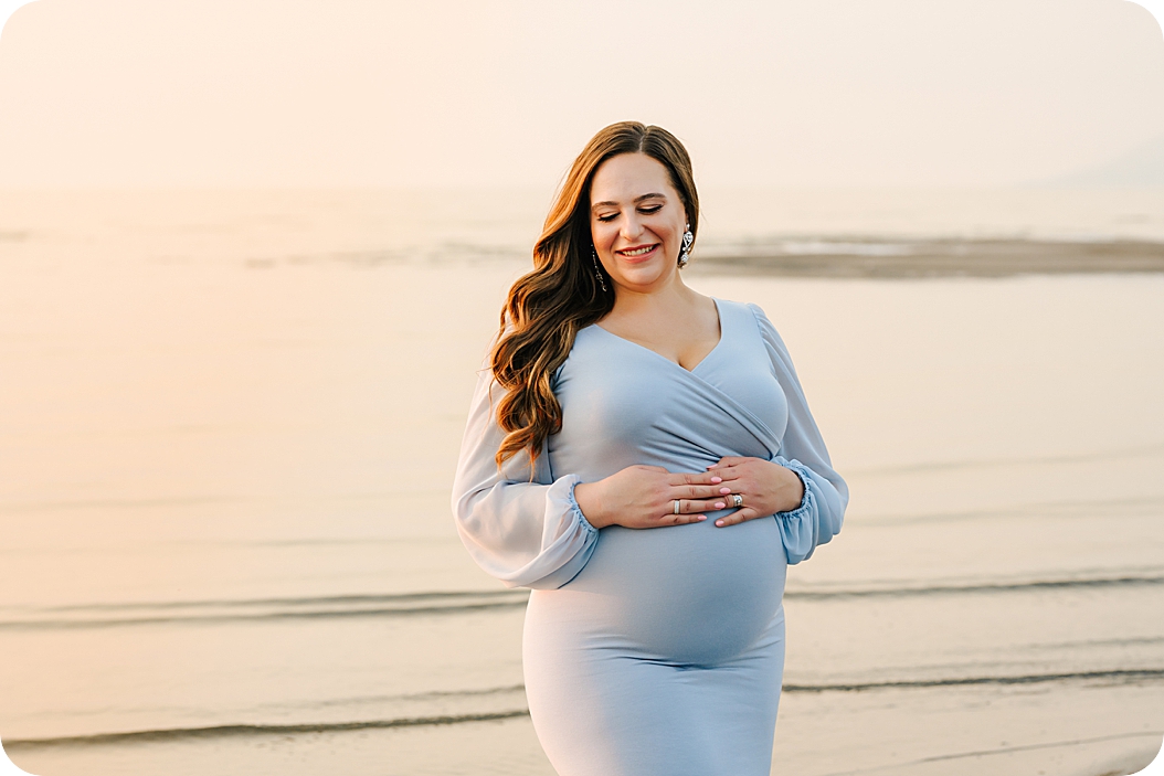 mom holds belly in blue dress during classic sunset maternity portraits on the lake