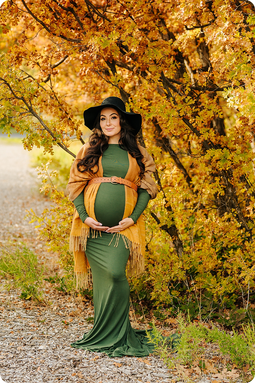 woman in wide brimmed hat holds belly in green gown during fall maternity portraits