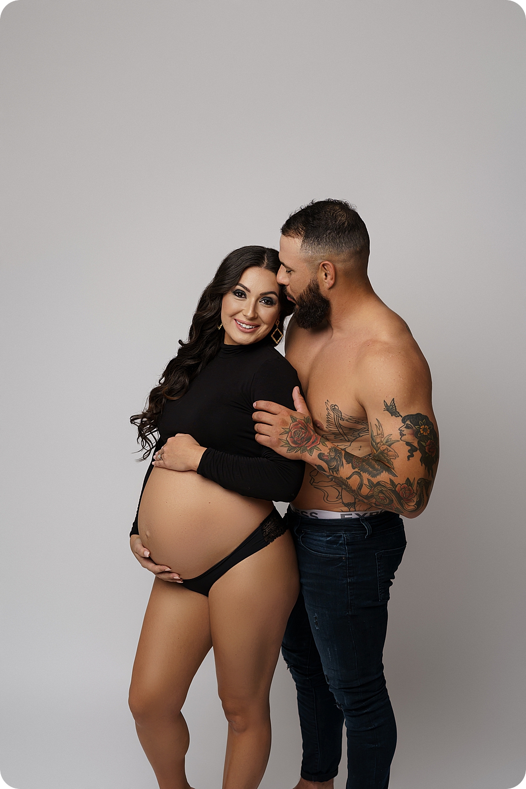 husband holds wife's arms while she holds belly during dramatic studio maternity portraits