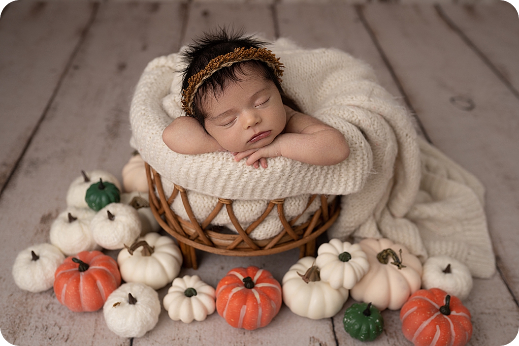 baby sleeps in basket by pumpkins for fall inspired newborn portraits 